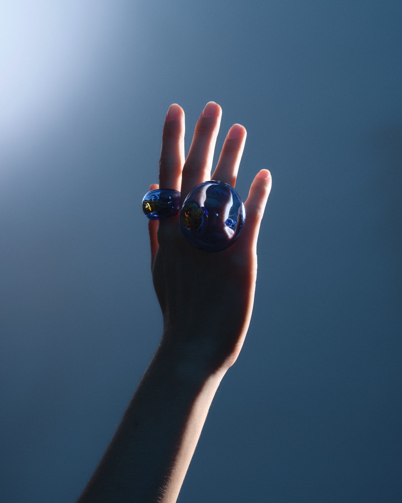 Atlantico ring handcrafted in light blue borosilicate glass, showcased on an upraised hand against a deep blue background, capturing the ring's translucent glow and sculptural design in dramatic lighting.