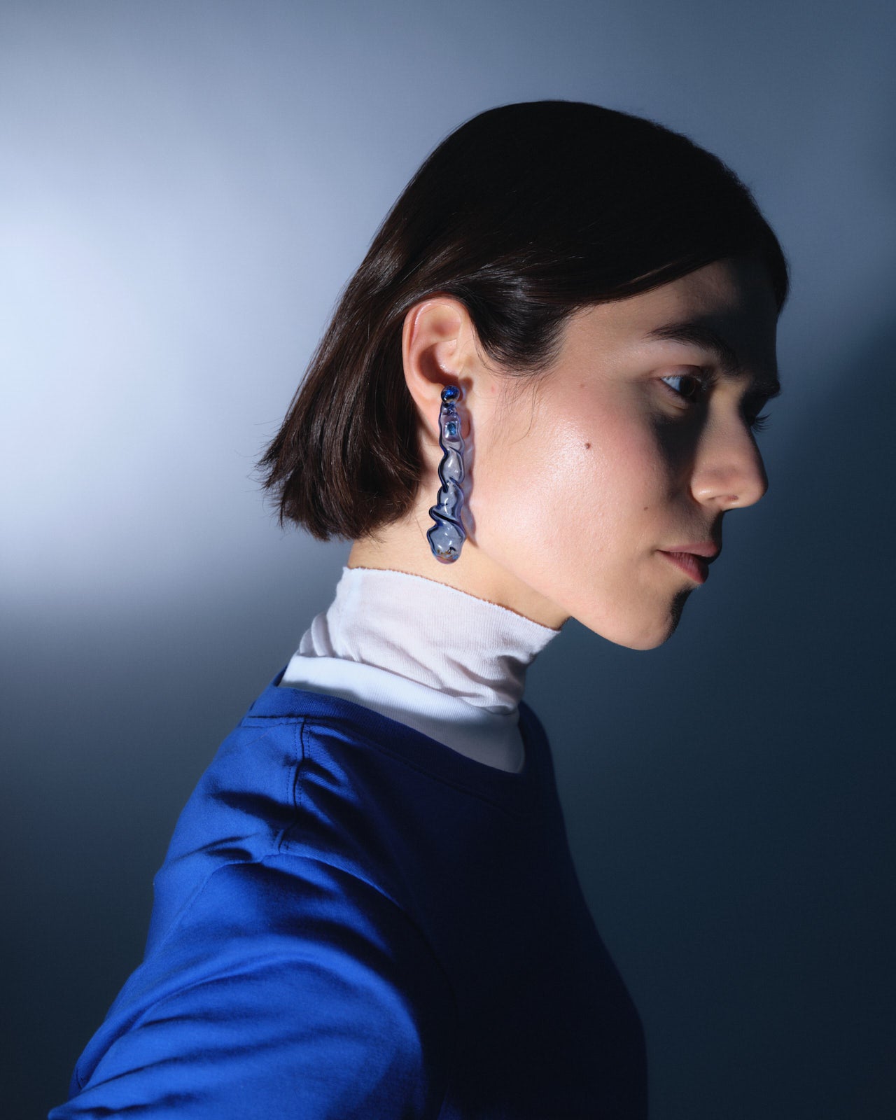 Side profile showcasing Indigo earrings, handcrafted borosilicate glass in deep blue, elegantly displayed against a navy and white outfit. The elongated 7cm sculptural design catches light dramatically against a dusky blue background.