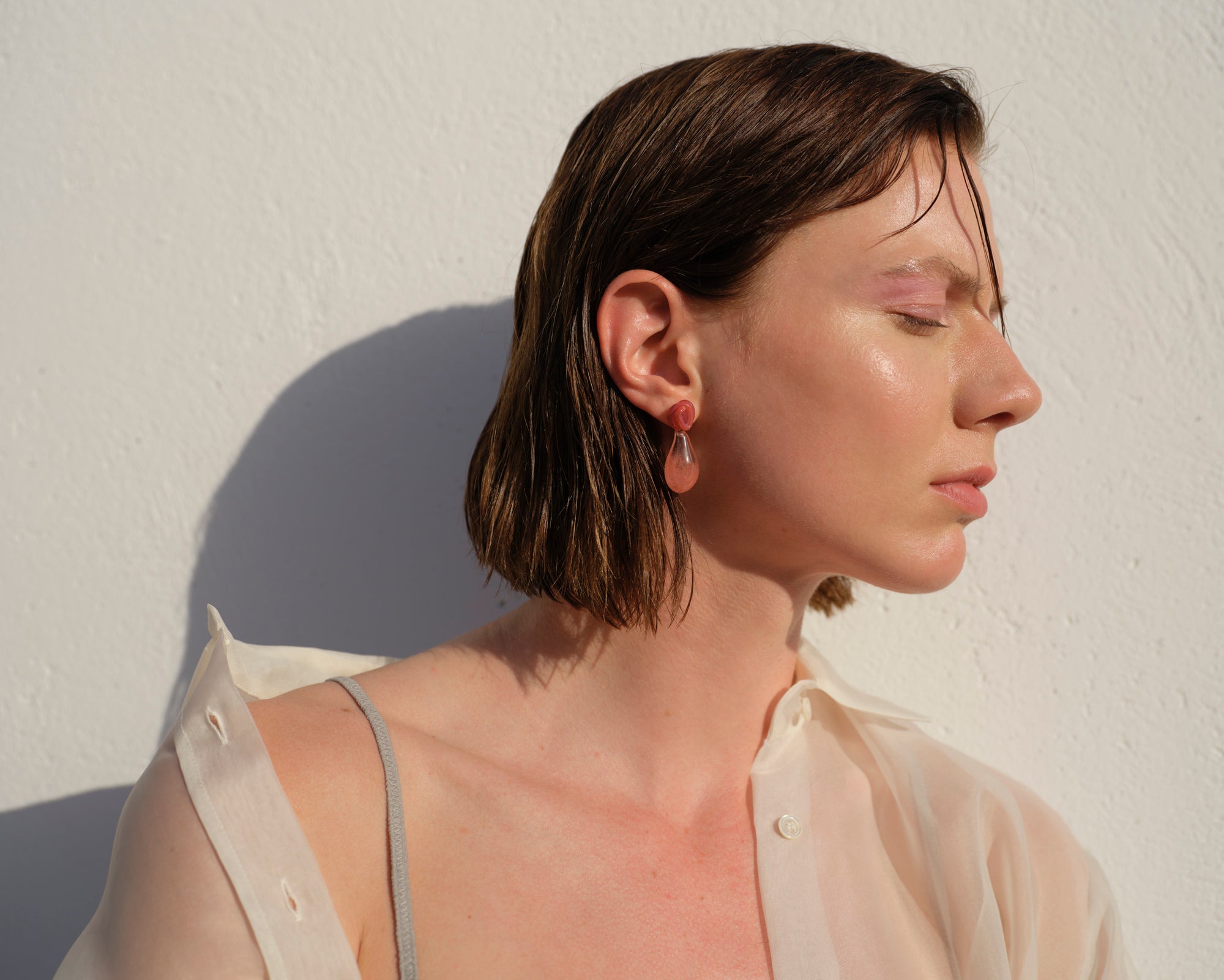 Handcrafted Lady in Red earrings shown in natural sunlight against white wall, featuring elegant red borosilicate glass drops measuring 3.5x1.5cm. Model wearing light cream shirt displaying side profile to showcase lightweight design and delicate craftsmanship from Barcelona.